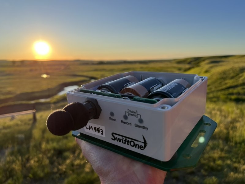 A SwiftOne acoustic recorder held up against a sunrise over grassland